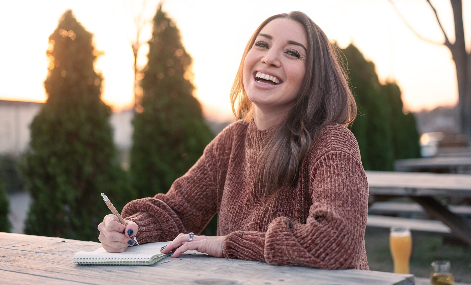 A young woman with long auburn hair, wearing a cozy brown sweater, smiles at the camera while writing in a spiral-bound notebook. She is seated at an outdoor wooden picnic table during sunset, with warm sunlight filtering through a row of tall evergreen trees in the background.